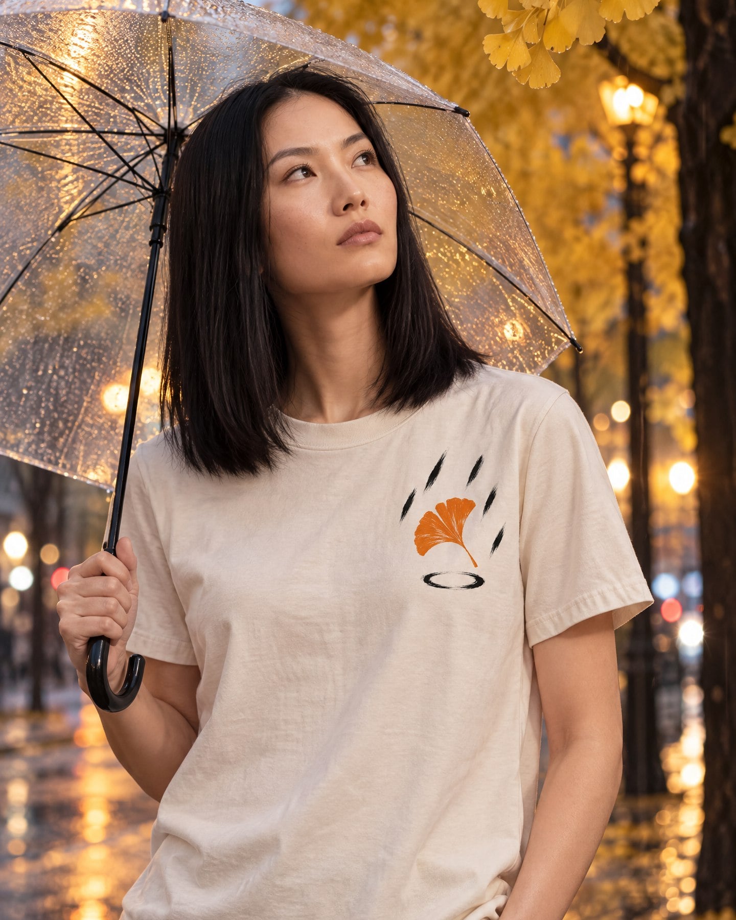 Photoreal image of an East Asian woman wearing the Ginkgo, Rain tee in Bone under golden ginkgo trees on a rain-wet street, holding a clear umbrella.