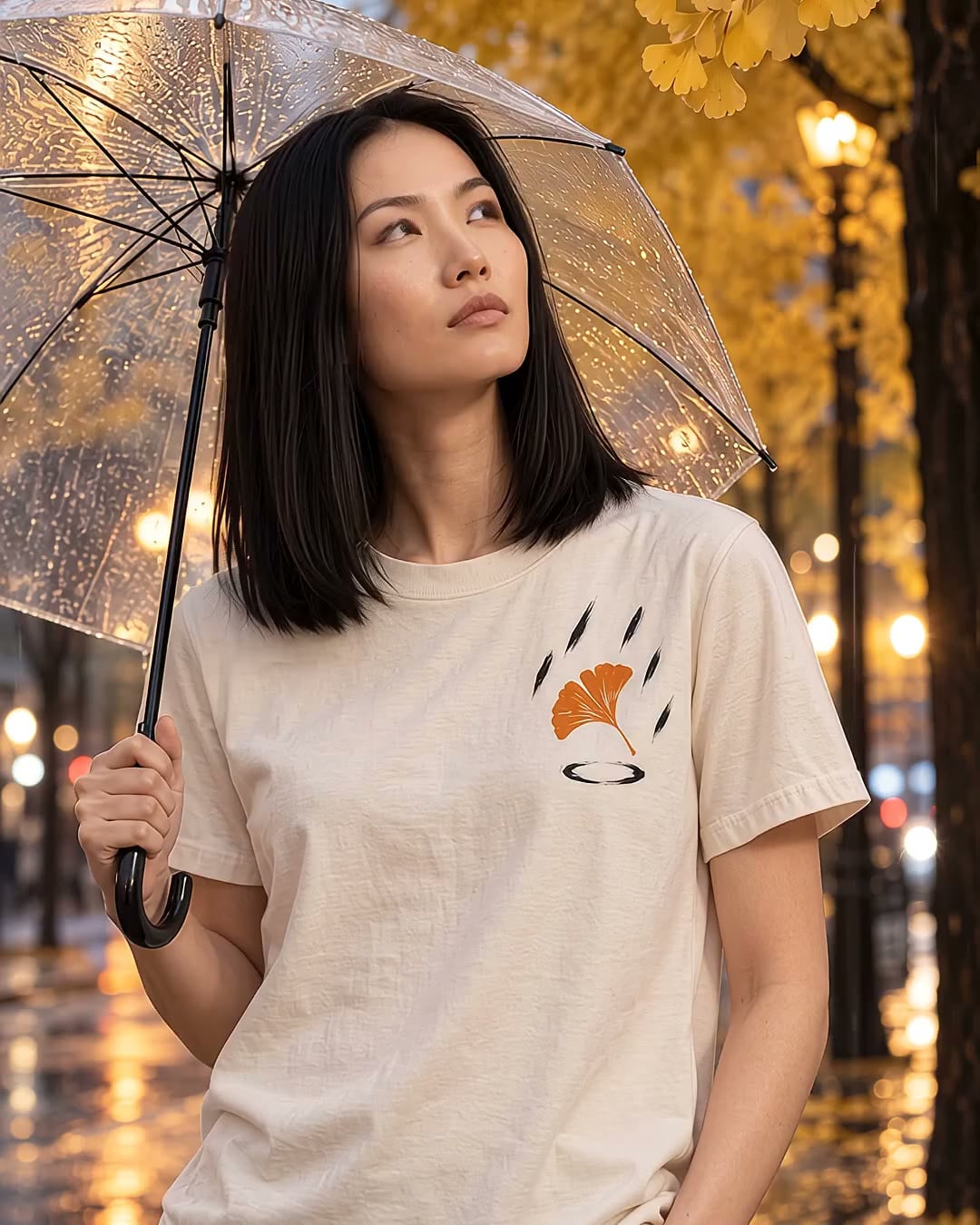The Ginkgo, Rain tee in Bone, laid flat, with a small print on the left chest: an orange ginkgo leaf falling through bold brushed rain strokes, with an ink ripple below.