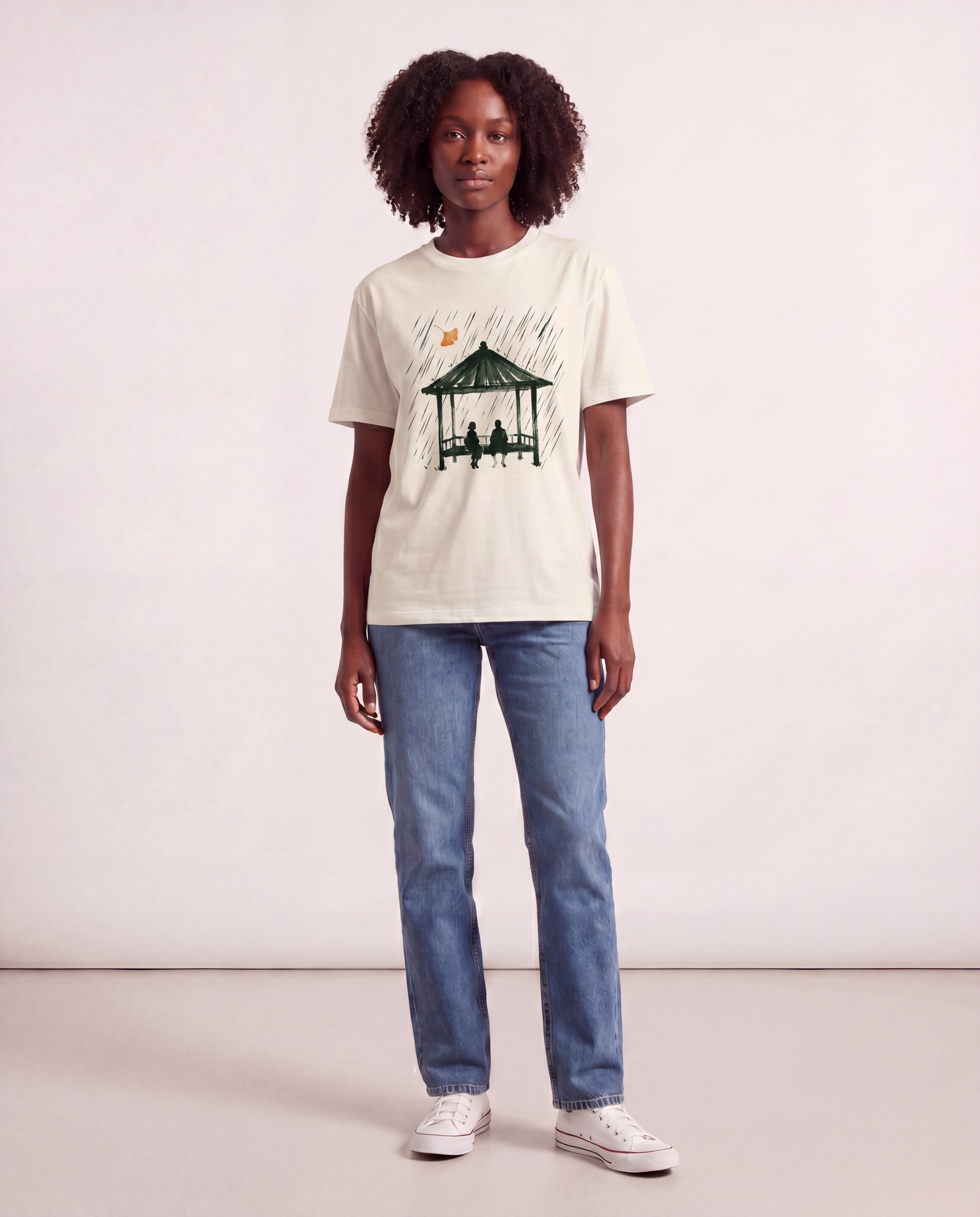 Editorial photograph of a Black female model wearing the Gazebo tee, full body, denim jeans and white sneakers, standing against a cream studio backdrop. LV-tier register.