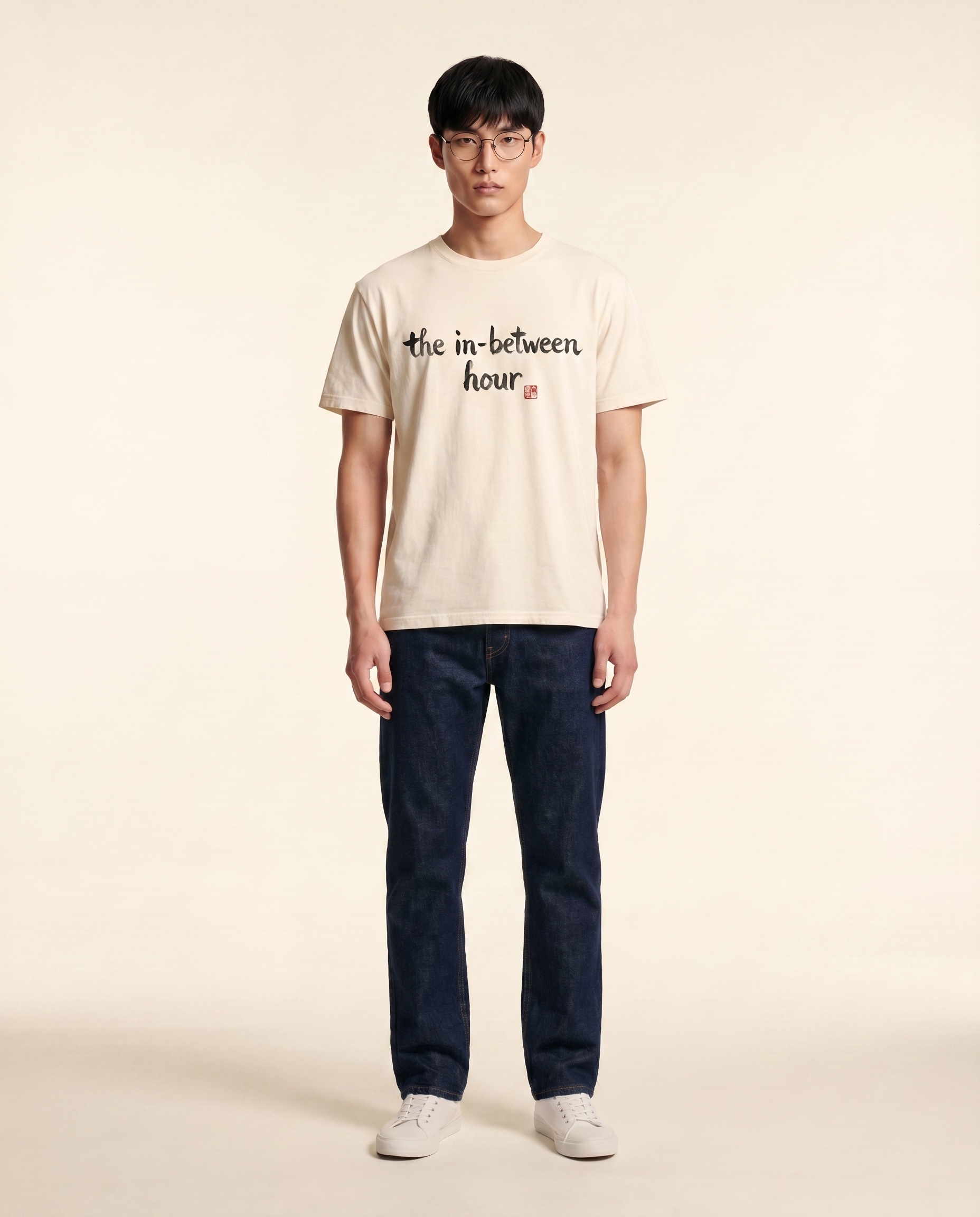 Editorial photograph of an East Asian male model with wire-frame glasses wearing the 'the in-between hour' tee, full body, dark indigo straight-leg denim jeans and white sneakers, standing against a cream studio backdrop. LV-tier register.