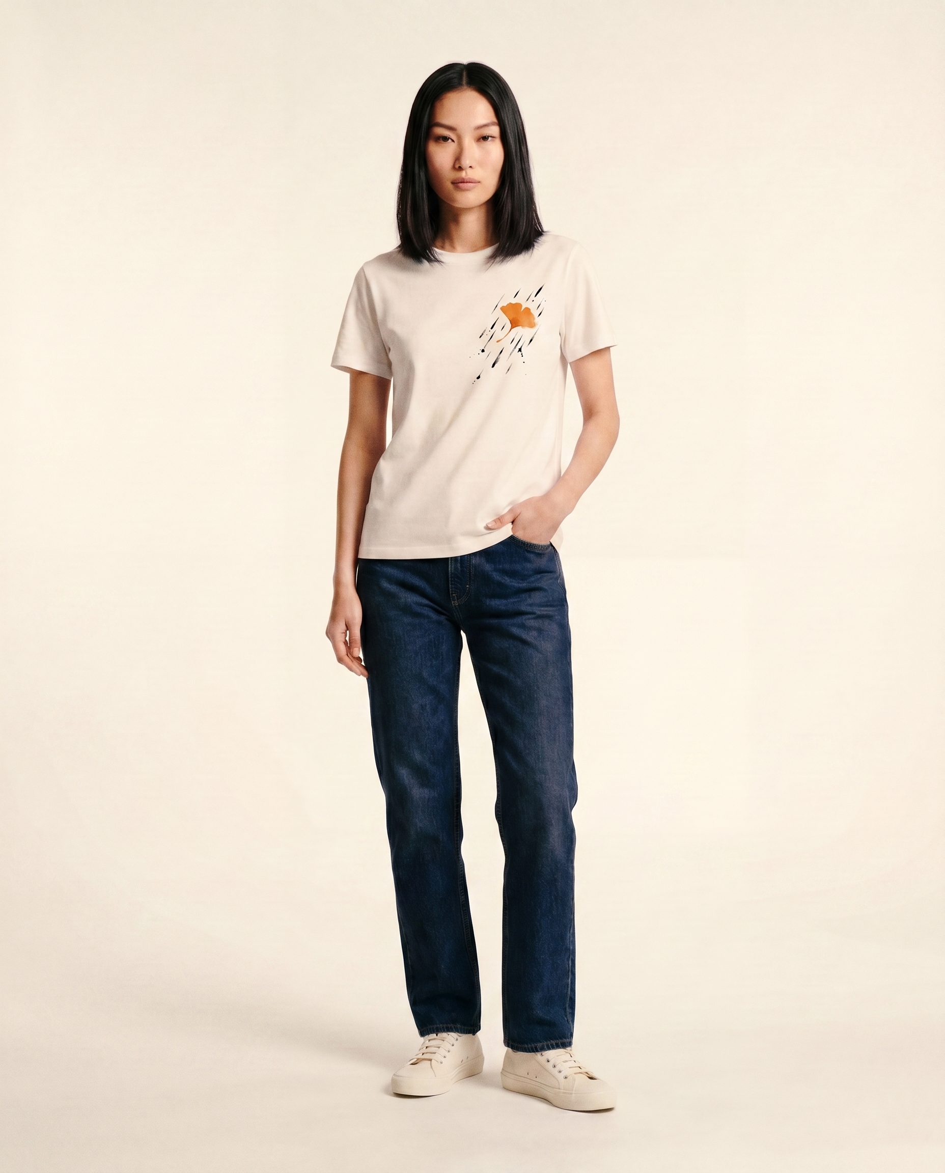 Editorial photograph of an East Asian female model wearing the Ginkgo, Rain tee, full body, dark indigo straight-leg denim jeans and white sneakers, standing against a cream studio backdrop. LV-tier register.