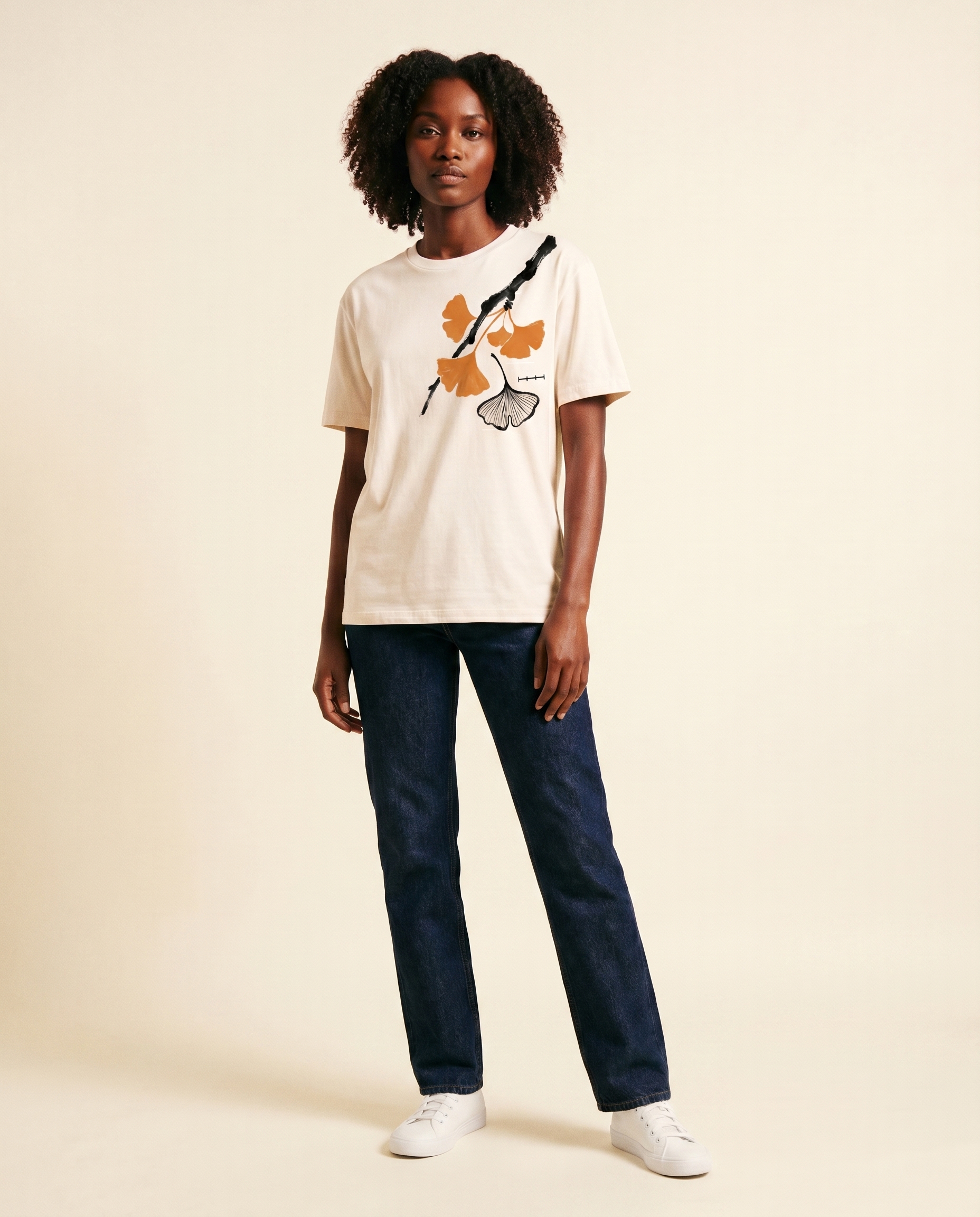 Editorial photograph of a Black female model with natural shoulder-length curls wearing the Ginkgo Study tee, full body, dark indigo straight-leg denim jeans and white sneakers, standing against a cream studio backdrop. LV-tier register.
